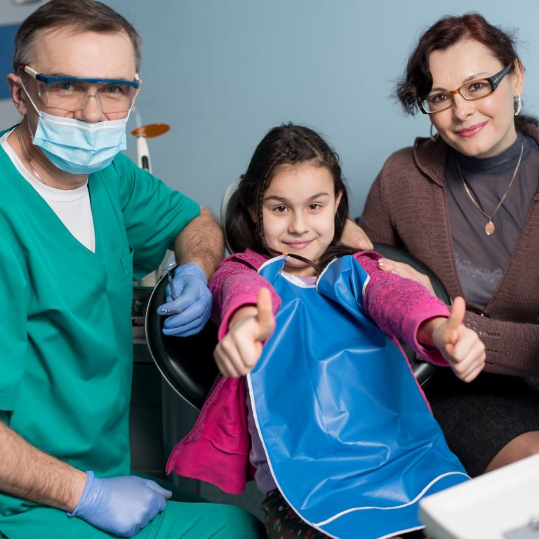 Portrait of senior pediatric dentist and girl with her mother on the first dental visit at the dental office. Young patient is smiling, showing thumbs up. Dentistry, medicine and health care concept