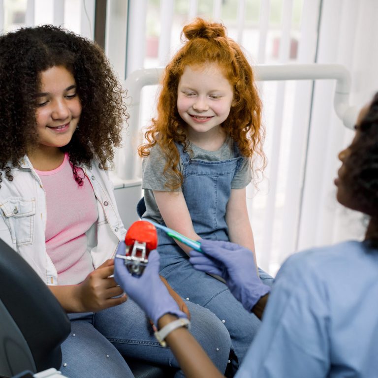 Pediatric dentistry. Back view of African lady dentist, showing how to brush teeth on jaw model, to her little patients, two smiling multiracial shcool girls, African American and Caucasian.